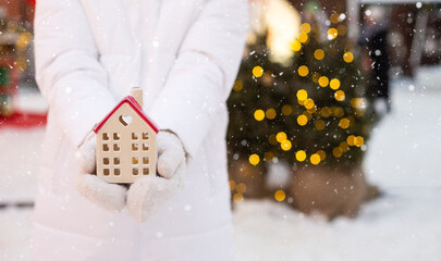 House, miniature cottage in hands of woman wearing mittens and warm clothes outdoor in snow. Deal for real estate, purchase, construction, relocation, mortgage. Cozy home, Christmas, new year booking