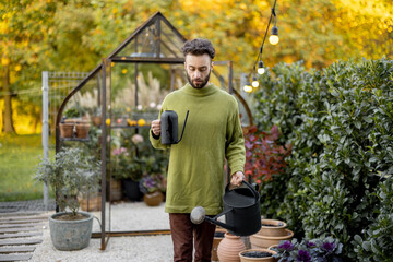 Fototapeta na wymiar Stylish guy watering plants, taking care of them in beautiful garden with small glass house on background. Hobby, work in garden concept