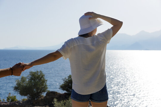 Portrait of a young woman by the sea, wearing a shirt and a hat and looking into the distance. An invisible man is holding her hand. Back photo. High quality photo
