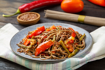 Soba with vegetables in soy sauce sprinkled with flax seeds in a plate on a napkin next to onions, tomatoes, peppers and flax seeds.
