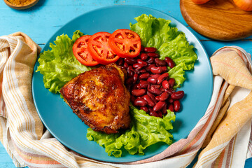 Chicken with red beans and tomato on lettuce in a blue plate on a blue table on a towel next to onions and spices.