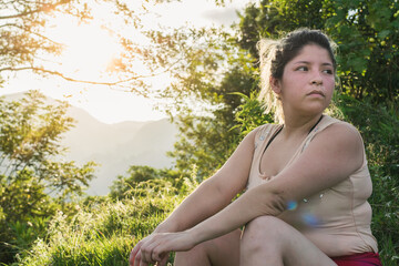 Naklejka na ściany i meble beautiful young latin woman sitting on a grassy meadow on the top of a mountain with a beautiful golden sunset in the background, curiously observing her surroundings.