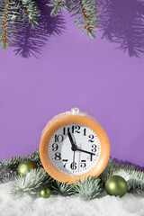 Fototapeta na wymiar Wood alarm clock with fir twigs and green baubles, balls, trinkets under snow. Purple paper background with copy-space. Traditional Xmas decor. Merry Christmas and Happy New Year.