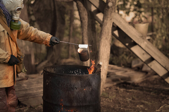 Post-apocalypse. A Man In A Gas Mask Holds A Tin Can Over A Fire Lit In A Rusty Iron Barrel. Selective Focus