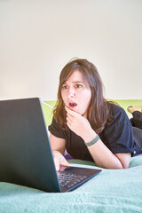 Vertical closeup of the amazed woman lying on the bed looking at the laptop.