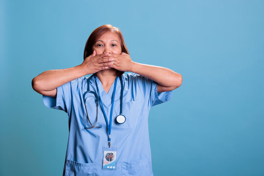 Elderly Physician Nurse In Blue Uniform Covering Mouth With Hands, Speak No Evil, Three Wise Monkeys Concept. Medical Assistant Working At Health Care Expertise Planning Disease Treatment