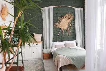 Modern boho interior of bedroom with green brick wall, bed, white wooden commode, curtains, houseplants, rattan basket, dray palm leaf and design personal accessories. Stylish home decor. Template. © Andriy Medvediuk
