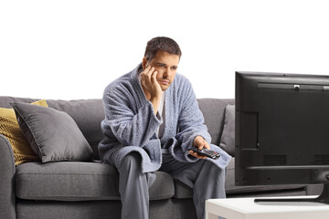 Bored young man in a bathrobe watching tv and sitting on a sofa