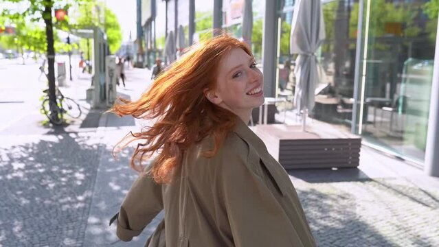 Cool teen stylish redhead fashion hipster girl model with red hair walking on big city street. Happy teenage girl with red hair wearing trench coat going in urban location along modern sunny avenue.