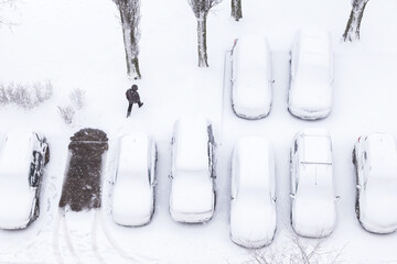 Snowfall, blizzard in town. Cars covered snow on a parking in blizzard, snowfall, top view. Man walk through snowdrift