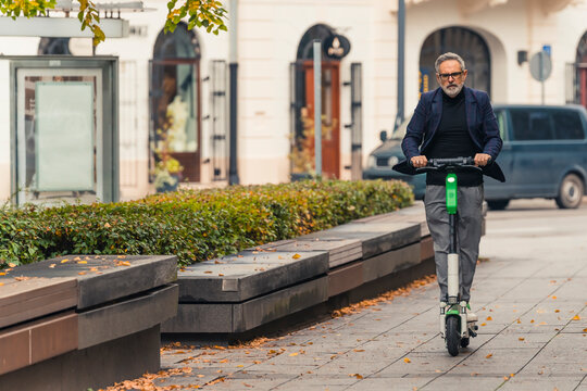 Grey-haired middle aged man in eyeglasses and suit riding rental electric scooter through city to work. Horizontal outdoor shot. High quality photo