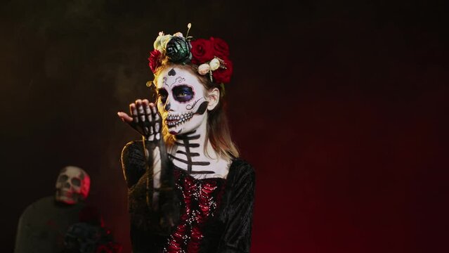 Creepy beautiful model sending air kisses on camera, wearing flowers crown and skull make up with kissy face. Looking like la cavalera catrina, being flirty on day of the dead. Handheld shot.