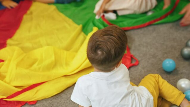Group of little children playing around on rainbow play parachute and throwing plastic balls. Multiracial nursery. Horizontal indoor video. High quality 4k footage
