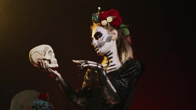 La cavalera catrina talking to skull in studio, acting scary and horror to celebrate mexican halloween tradition. Santa muerte dressed in horror spooky costume with body art. Handheld shot.