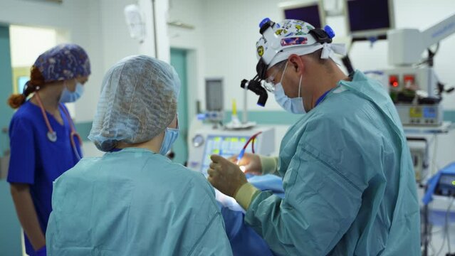 Medics performing surgery standing their backs to camera. Surgeon inserts the instrument into patient's nose. Diverse equipment at backdrop.