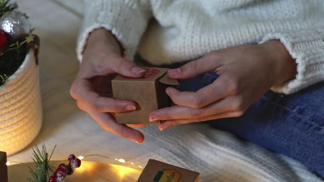 Hands of a caucasian teenage girl open an empty kraft advent calendar box getting ready to put a gift in it, sitting on the bed, side view close-up in slow motion.
