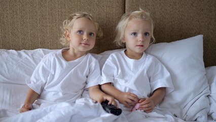 Little twin sisters sit in bed leaning on pillows. They are watching TV which hangs on the wall. The screen shows children's content. From time to time they change channels.