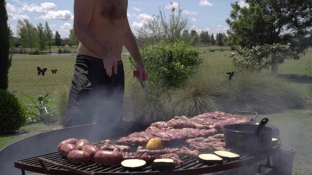 Barbecue outdoor. View of a caucasian man roasting veal, beef, pork sausages, blood sausages, ribs, chitterlings and other meat cuts, in the iron grill with a smokey fire.