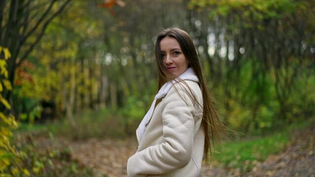Long-haired brunette woman standing in the autumn park turns around to the camera. Lady looks into camera slowly approaching to her face. Blurred backdrop.
