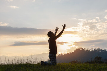 Young male kneeling down with hands open palm up praying to God on the mountain sunset background. - obrazy, fototapety, plakaty