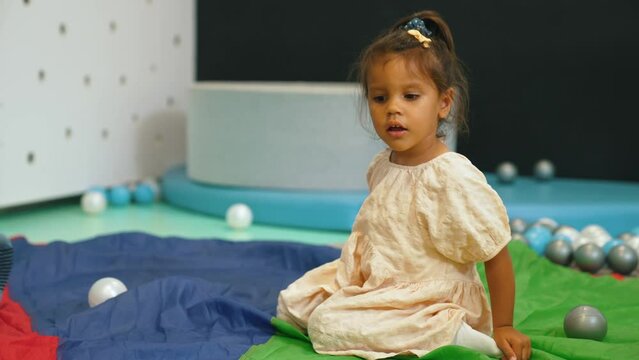 Little dark-skinned girl in nursery sitting on rainbow play parachute looking around with curious expression and kids throwing balls in the background. Horizontal indoor video . High quality 4k