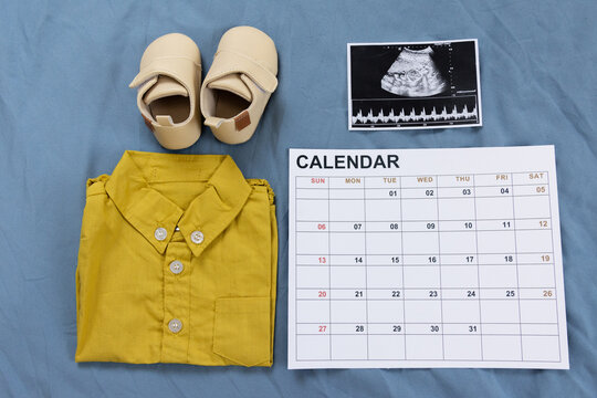 yellow newborn clothes, cream color shoes, ultrasound image and calendar on blue background. concept of pregnancy and giving birth