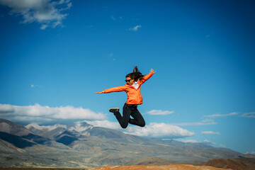 Young female traveller jumping with hands up against the mountains and blue sky. Happy woman enjoying active vacation. Travel and fun concept.