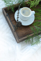 Cup of black morning coffee and green branches and forest cones on a wooden tray on white bedroom