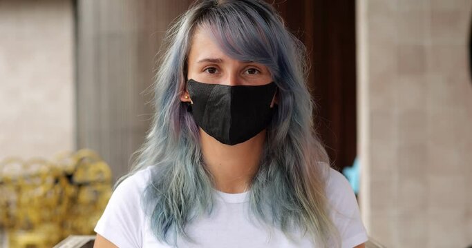 Portrait shot of young woman wearing black protective mask, blurred background. Hipster girl with dyed long hair looking straight to camera, not a joyful and concentrated expression on her face