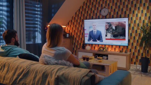 Couple sitting on couch in living room with snacks, watching TV news about military exercises, NATO training, armed forces. Man and woman spending evening at home. TV live broadcasting.