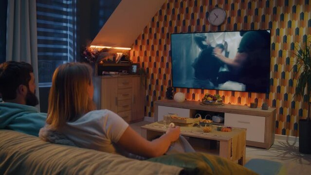 Happy couple sitting on sofa, watching action movie on TV or criminal blockbuster on streaming service, talking and discussing acting, eating snacks, pizza, chips. Spouses resting at home in evening.