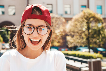 Young beautiful smiling hipster woman in trendy summer clothes. Sexy carefree woman posing on the street background in cap at sunset. Positive model outdoors. Cheerful and happy in sunglasses