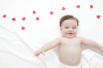top view of beautiful and smiling five month old baby boy lying on the bed covered with a white sheet and with red heart decorations.concept of Valentine's Day 