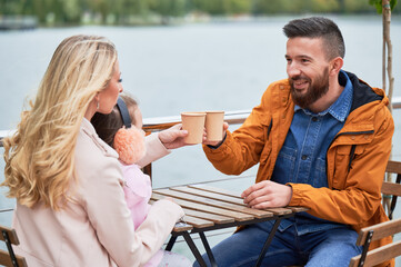 Cheerful man and woman toasting with cups of coffee while spending time with daughter in outdoor cafe. Happy husband and wife sitting at the table and clinking drink cups in new urban district.