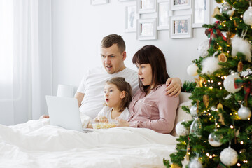 christmas, family and love concept - surprised family sitting on the bed and watching movie near christmas tree