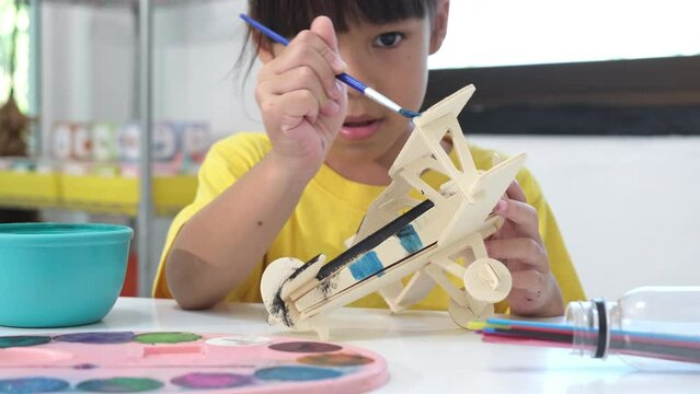 Portrait of a happy Asian girl with a brush painting on a wooden toy plane in the classroom. Arts and crafts for kids. Creative little artist at work.