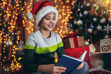 Obraz na płótnie Canvas Happy boy lying on front on floor by christmas tree, reading book surrounded by gifts.