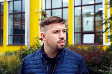 Looking away young caucasian 34 years old man in blue jacket with goatee beard on yellow facade of college modern building. Young university teacher and scientist. Head shot man lifestyle portrait. - obrazy, fototapety, plakaty