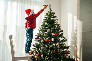 A boy in a santa claus hat decorates the New Year tree at home, puts a Christmas toy on the top of the tree. Children are preparing to celebrate Christmas and New Year.