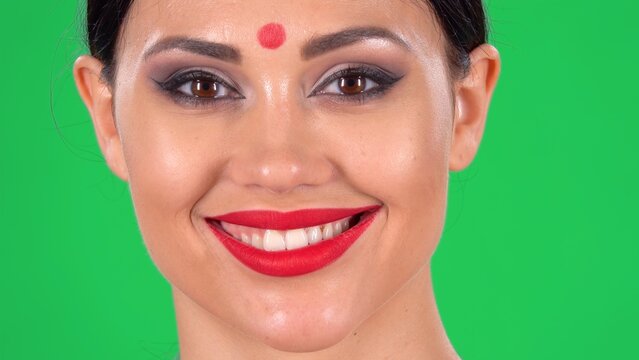 Portrait Of Beautiful Indian Woman With Red Dot On Forehead Looking At Camera And Smiling Revealing Teeth. Close Up Of A Brunettes Face With Bright Eye Makeup And Red Lips On Green Screen At Studio.