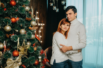 couple, a man and woman in the same white sweaters are hugging near a christmas tree..