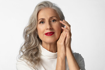 An elderly gray-haired attractive woman with bright red lips in a white stylish turtleneck poses with her hands near her face with a confident look