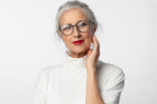 A lovely elderly intelligent woman with glasses and a slight smile touches her face with her hand. A cosmetic conceptual portrait of aging skin care.