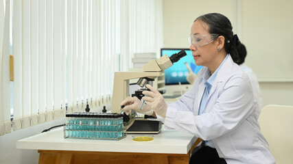 Mature female biotechnology specialist looking under microscope, conducting experiment in a laboratory