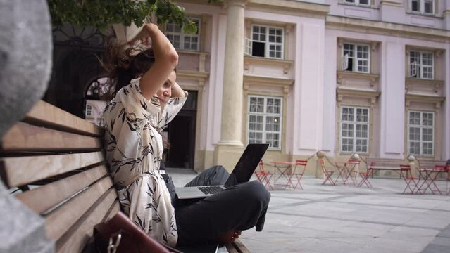 Young woman working on laptop on city bench in historical surroundings.