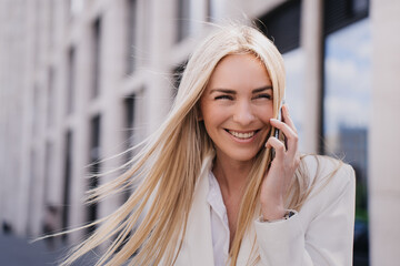Close up of blonde Italian woman with flattering loose hair talking by phone outside over blurry business center. Happy European female received great news. Student toothy smiling. Successful people.