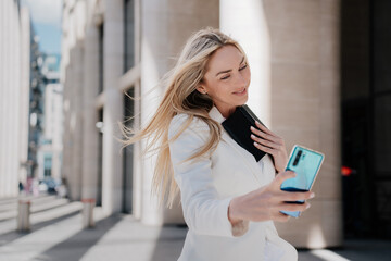 Gorgeous blonde woman in white suit walking by street smiling making selfie, talking by video call at summer day, going to work. Cheerful Italian successful businesswoman with flattering loose hair.