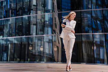 Exhausted young blonde woman in white suit standing outside over glassy building. Upset employee feels bad, loosing job, failure. Lonely businesswoman in depression hugging herself holding diary.