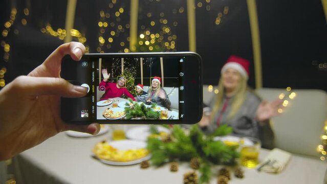 Close-up of the phone in the hands of a girl taking pictures of a family with a child for the New Year. Happy family waving his hands at camera during christmas party at