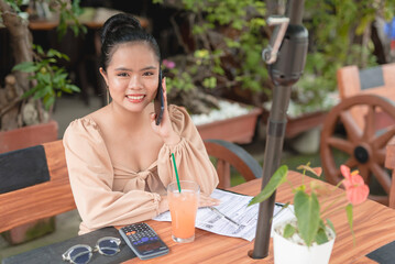 A smiling young woman sitting outside a fancy cafe is glad to hear the good news while talking on...